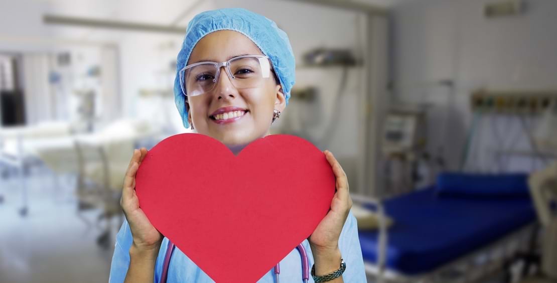 Unge pige i sygeplejeuniform holder et stort rødt hjerte foran sig og smiler ind i kameraet.